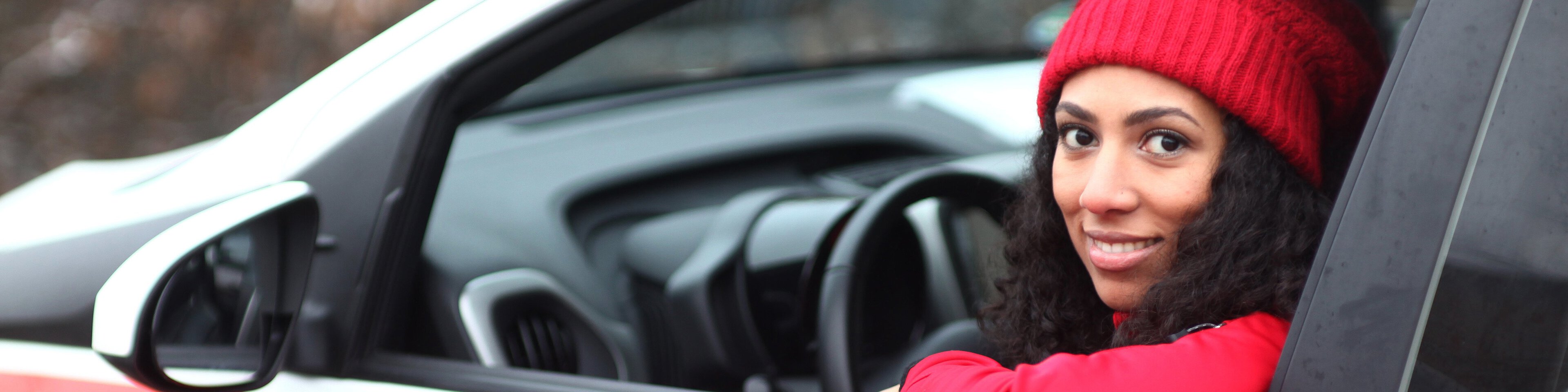 Dunkelhaarige Frau mit langen locken und roter Mütze und Pulli schaut lächelnd aus der Fahrertür eines Caritas Pflege Auto heraus. | © heydenaber